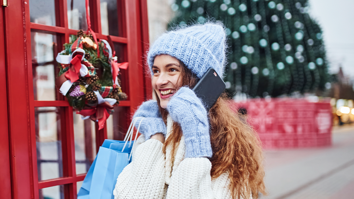 A smiling woman wearing winter clothes talks on her phone while shopping during the festive season, representing increased holiday customer service activity.